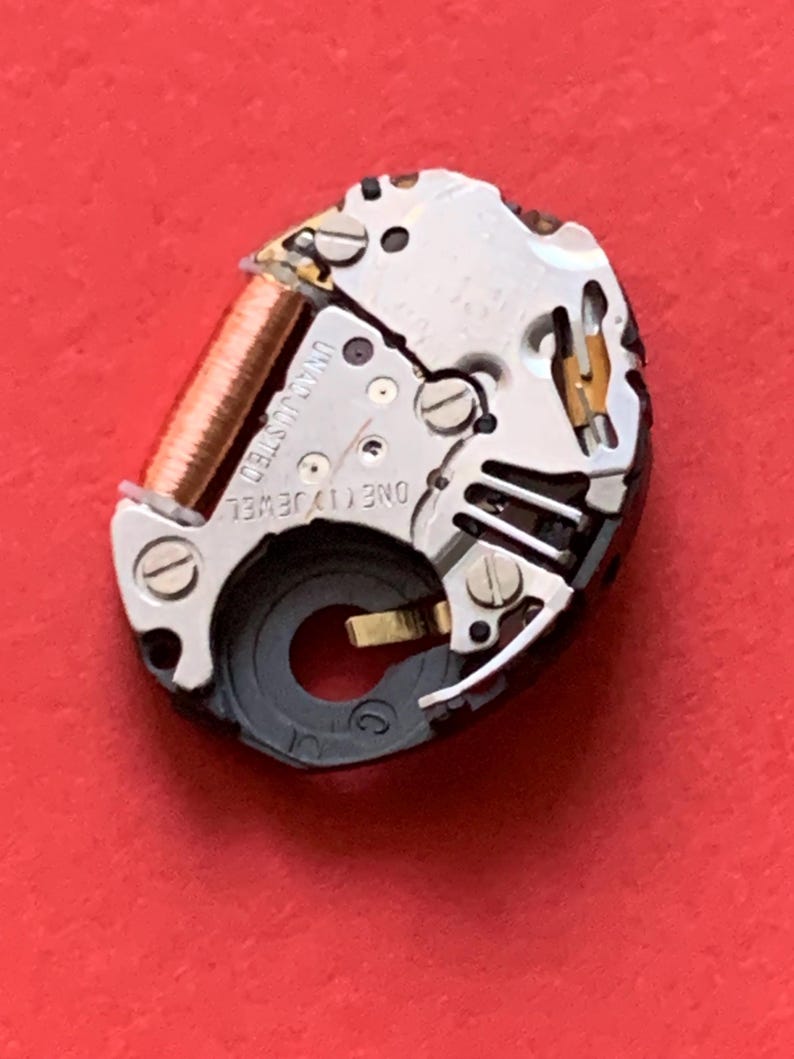 May include: Close-up of a watch movement with intricate gears, screws, and a copper coil. The words "UNADJUSTED" and "ONE (1) JEWEL" are visible. The watch mechanism is set against a vibrant red background.
