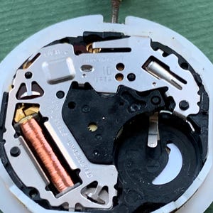 K&ouml;nnte beinhalten: Nahaufnahme eines Uhrwerks mit komplexen mechanischen Komponenten. Das Uhrwerk zeigt eine Kupferspule, silberfarbene Metallplatten und ein schwarzes Batteriefach. Der Hintergrund ist gr&uuml;n.