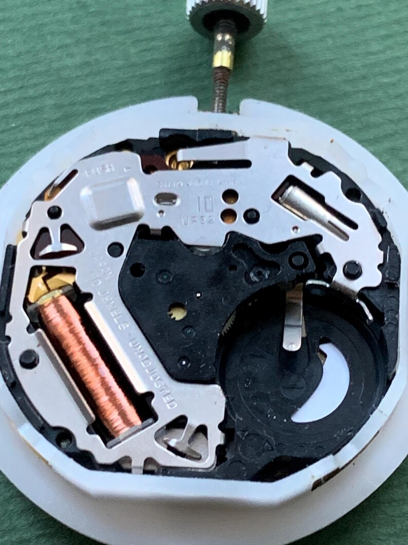 K&ouml;nnte beinhalten: Nahaufnahme eines Uhrwerks mit komplexen mechanischen Komponenten. Das Uhrwerk zeigt eine Kupferspule, silberfarbene Metallplatten und ein schwarzes Batteriefach. Der Hintergrund ist gr&uuml;n.