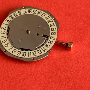 K&ouml;nnte beinhalten: Ein Uhrwerk mit einem wei&szlig;en Datumsring, der die Zahlen 1-31 anzeigt. Der silberfarbene Mechanismus hat eine kleine goldfarbene Krone und ist vor einem roten Hintergrund platziert. Das Uhrwerk ist f&uuml;r Reparatur oder Ersatz gedacht.