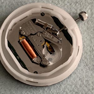 Puede incluir: Primer plano de un movimiento de reloj, alojado en una carcasa de pl&aacute;stico blanco. El mecanismo interno presenta una bobina de cobre, componentes met&aacute;licos plateados y un peque&ntilde;o tornillo. El texto "UNADJUSTED MIYOTA CO. 2115 JAPAN" es visible.
