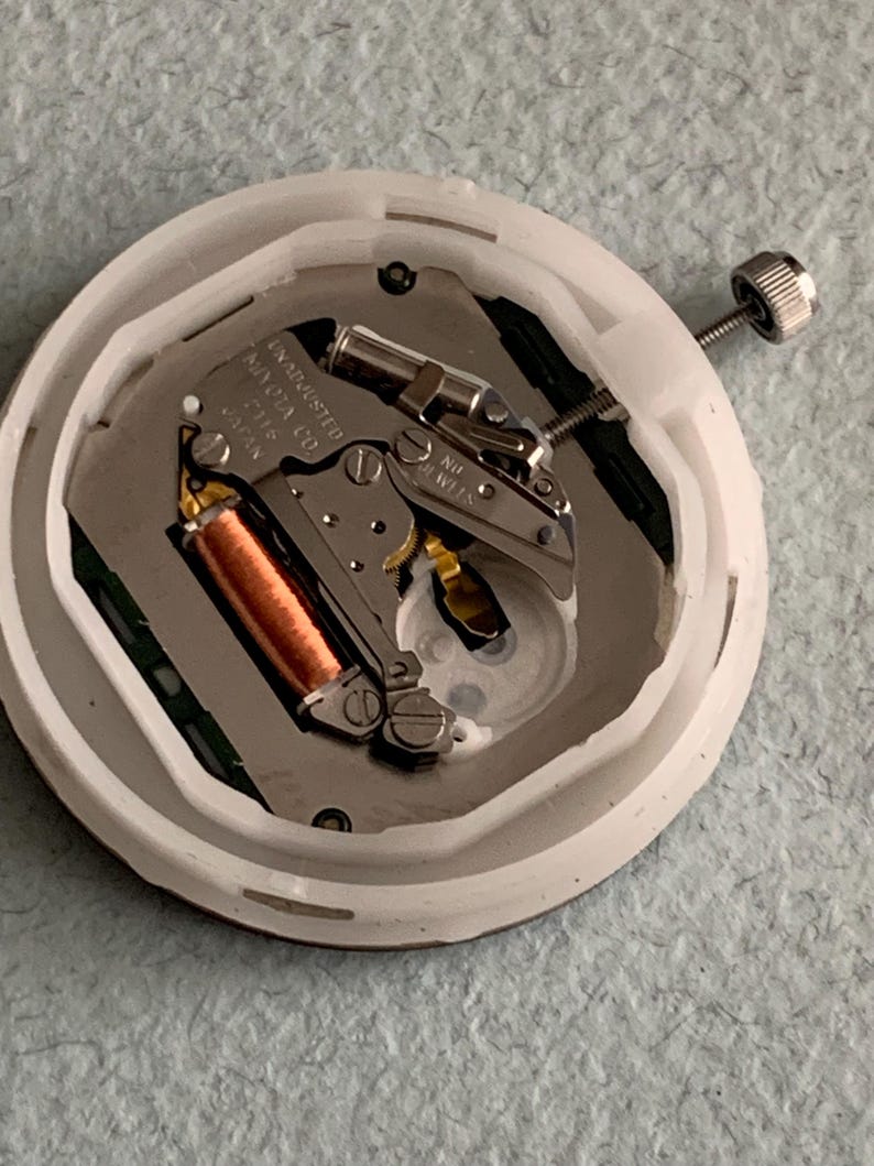 Puede incluir: Primer plano de un movimiento de reloj, alojado en una carcasa de pl&aacute;stico blanco. El mecanismo interno presenta una bobina de cobre, componentes met&aacute;licos plateados y un peque&ntilde;o tornillo. El texto "UNADJUSTED MIYOTA CO. 2115 JAPAN" es visible.
