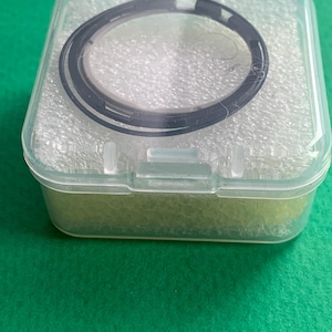 May include: A black circular object, possibly a watch component, is encased in a clear plastic box. The box is rectangular with a hinged lid and sits on a green surface. The object has a circular design with a dark outer ring.