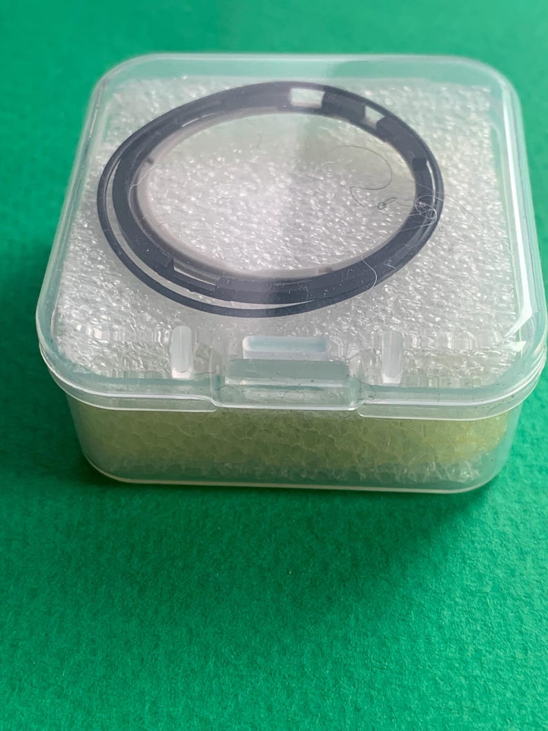 May include: A black circular object, possibly a watch component, is encased in a clear plastic box. The box is rectangular with a hinged lid and sits on a green surface. The object has a circular design with a dark outer ring.