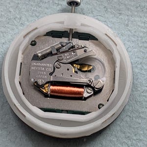 Puede incluir: Un movimiento de reloj con el texto "UNADJUSTED MIYOTA CO. 2115 JAPAN" es visible dentro de una carcasa de pl&aacute;stico blanco. El movimiento presenta varios componentes met&aacute;licos, incluyendo una bobina de cobre y un volante plateado. Un v&aacute;stago plateado se extiende desde la parte superior.