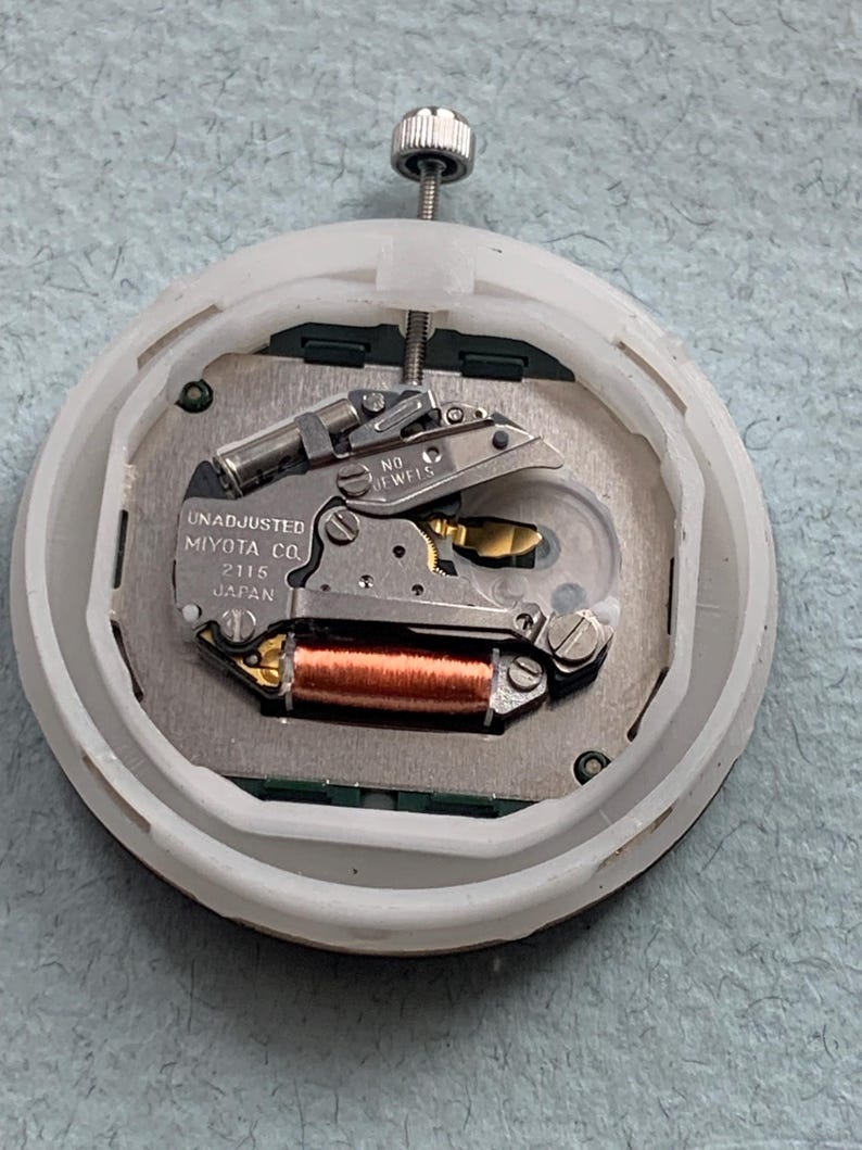 Puede incluir: Un movimiento de reloj con el texto "UNADJUSTED MIYOTA CO. 2115 JAPAN" es visible dentro de una carcasa de pl&aacute;stico blanco. El movimiento presenta varios componentes met&aacute;licos, incluyendo una bobina de cobre y un volante plateado. Un v&aacute;stago plateado se extiende desde la parte superior.
