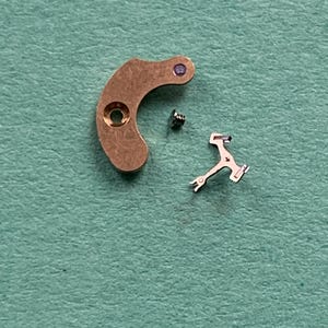 May include: Close-up of watch parts, including a curved, copper-colored lever with a hole and a purple jewel, a small screw, and a silver-colored escapement fork. The parts are arranged on a textured, teal-colored surface.