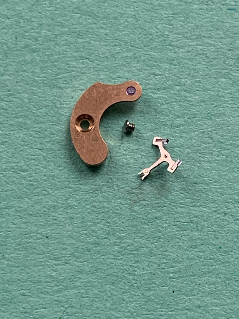 May include: Close-up of watch parts, including a curved, copper-colored lever with a hole and a purple jewel, a small screw, and a silver-colored escapement fork. The parts are arranged on a textured, teal-colored surface.