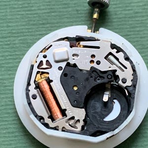 K&ouml;nnte beinhalten: Nahaufnahme eines Uhrwerks mit komplexen mechanischen Komponenten. Der Uhrenmechanismus verf&uuml;gt &uuml;ber silberfarbene Metallteile, eine kupferfarbene Spule und eine kleine Batterie. Oben ist eine silberne Krone befestigt. Das Uhrwerk befindet sich auf einer wei&szlig;en, kreisf&ouml;rmigen Basis.