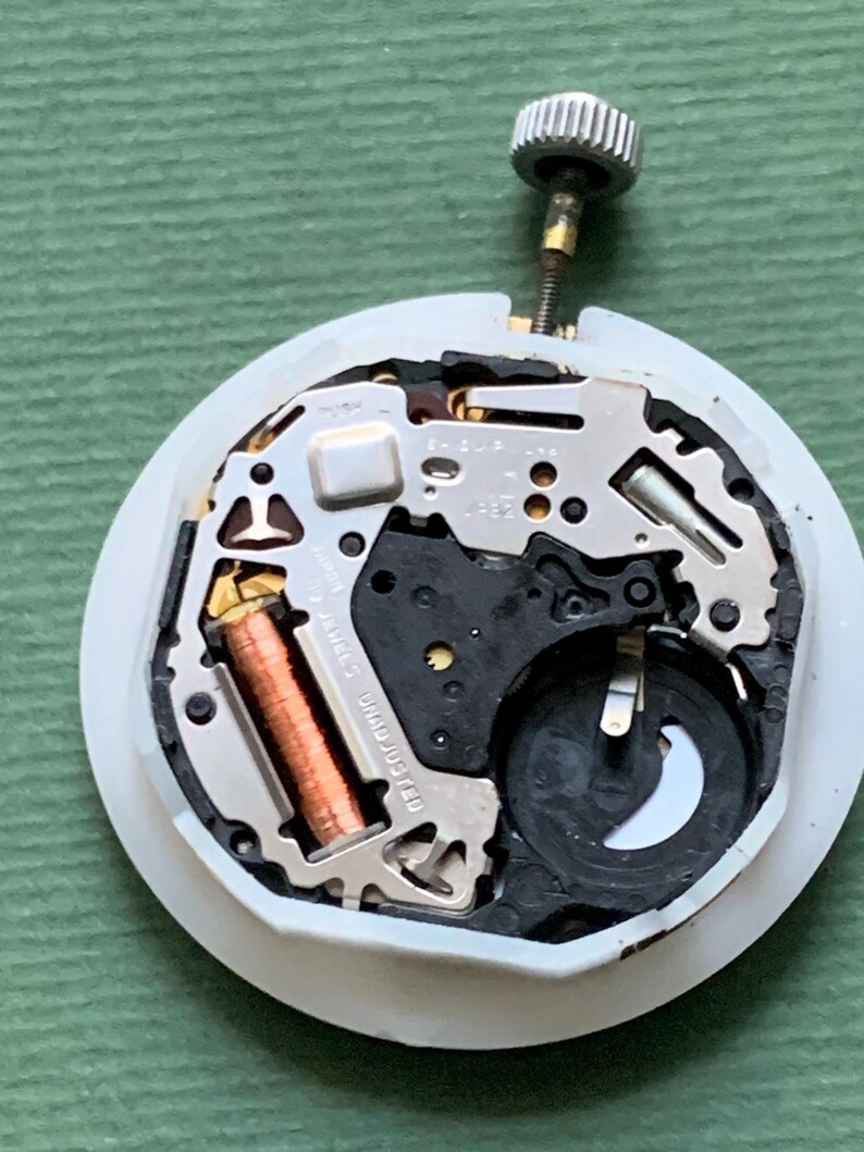 K&ouml;nnte beinhalten: Nahaufnahme eines Uhrwerks mit komplexen mechanischen Komponenten. Der Uhrenmechanismus verf&uuml;gt &uuml;ber silberfarbene Metallteile, eine kupferfarbene Spule und eine kleine Batterie. Oben ist eine silberne Krone befestigt. Das Uhrwerk befindet sich auf einer wei&szlig;en, kreisf&ouml;rmigen Basis.