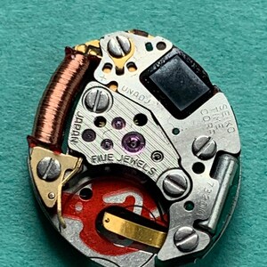 May include: Close-up of a watch movement with the text "FIVE JEWELS" and "SEIKO TIME CORP". The movement has a copper coil, silver-colored metal components, and a red painted section. The background is a solid teal color.