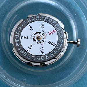 May include: A watch movement with a white dial showing days of the week and numbered dates. It has a silver outer ring and a small crown. The text "U-RING" is visible.
