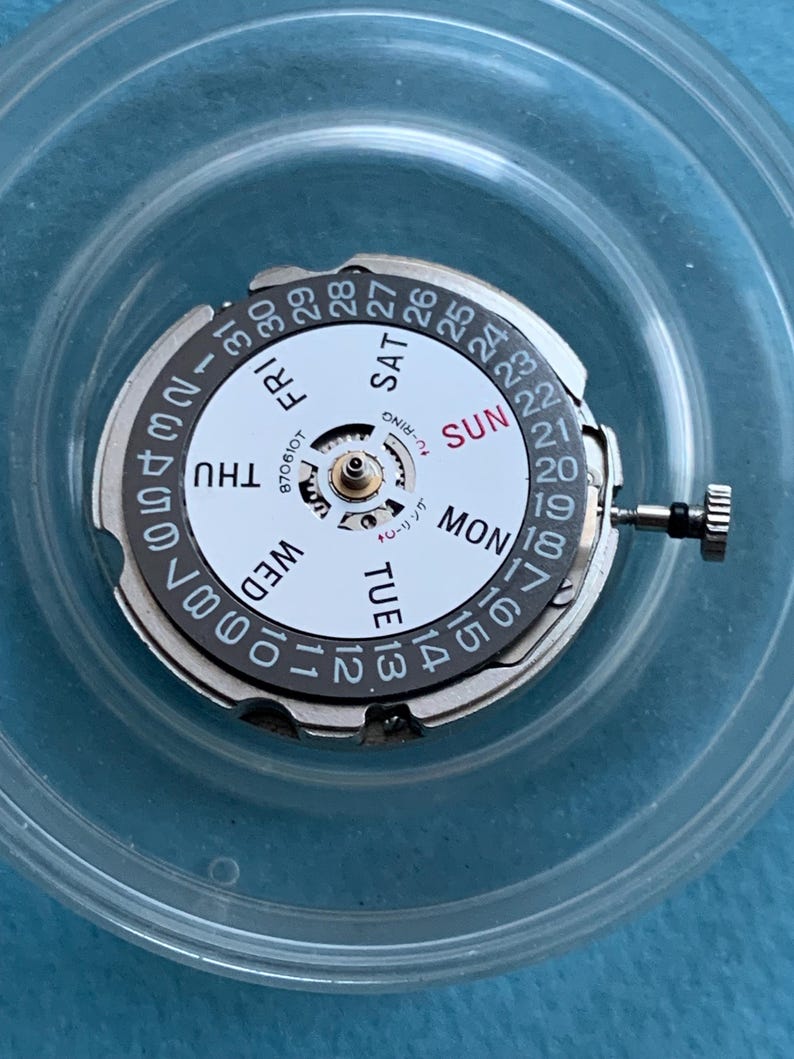 May include: A watch movement with a white dial showing days of the week and numbered dates. It has a silver outer ring and a small crown. The text "U-RING" is visible.