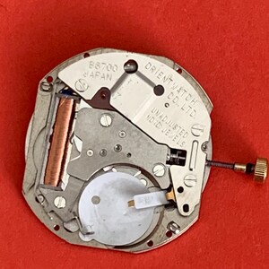 K&ouml;nnte beinhalten: Nahaufnahme eines Uhrwerks mit der Aufschrift "ORIENT WATCH CO., LTD." und "UNADJUSTED". Das Uhrwerk ist silberfarben mit Kupfer- und Goldakzenten, vor rotem Hintergrund.
