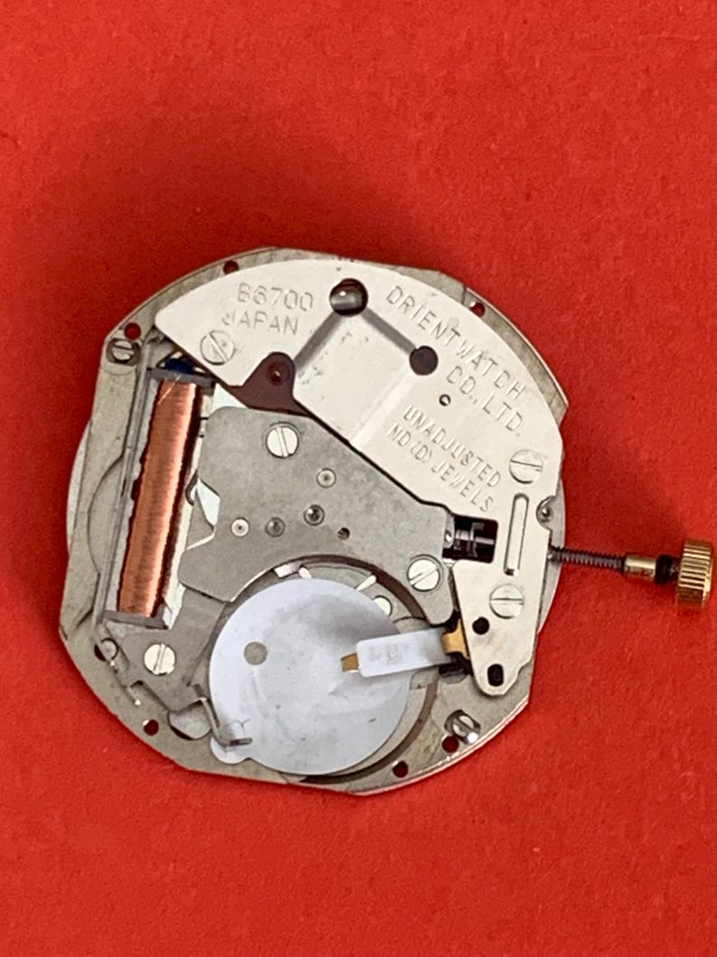 K&ouml;nnte beinhalten: Nahaufnahme eines Uhrwerks mit der Aufschrift "ORIENT WATCH CO., LTD." und "UNADJUSTED". Das Uhrwerk ist silberfarben mit Kupfer- und Goldakzenten, vor rotem Hintergrund.