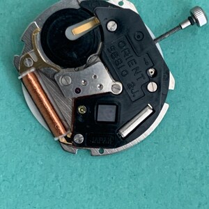 May include: A close-up of a watch movement with the text "ORIENT 58810 2J" and "JAPAN" visible. The movement has a silver metal frame, a black plastic component, and a copper coil. A small screw is attached to the side.