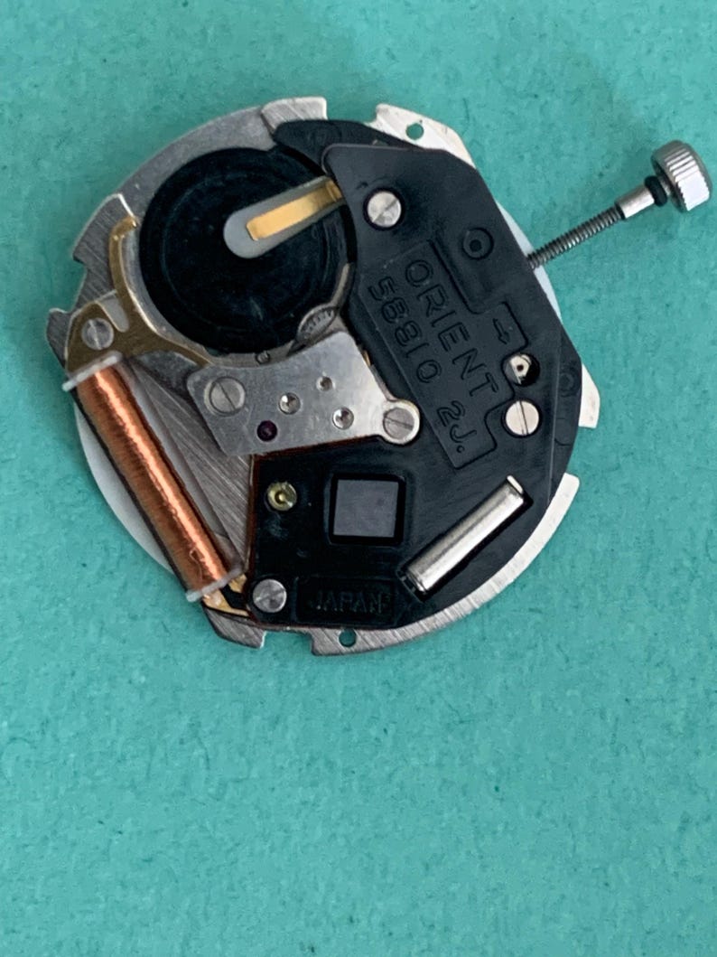 May include: A close-up of a watch movement with the text "ORIENT 58810 2J" and "JAPAN" visible. The movement has a silver metal frame, a black plastic component, and a copper coil. A small screw is attached to the side.
