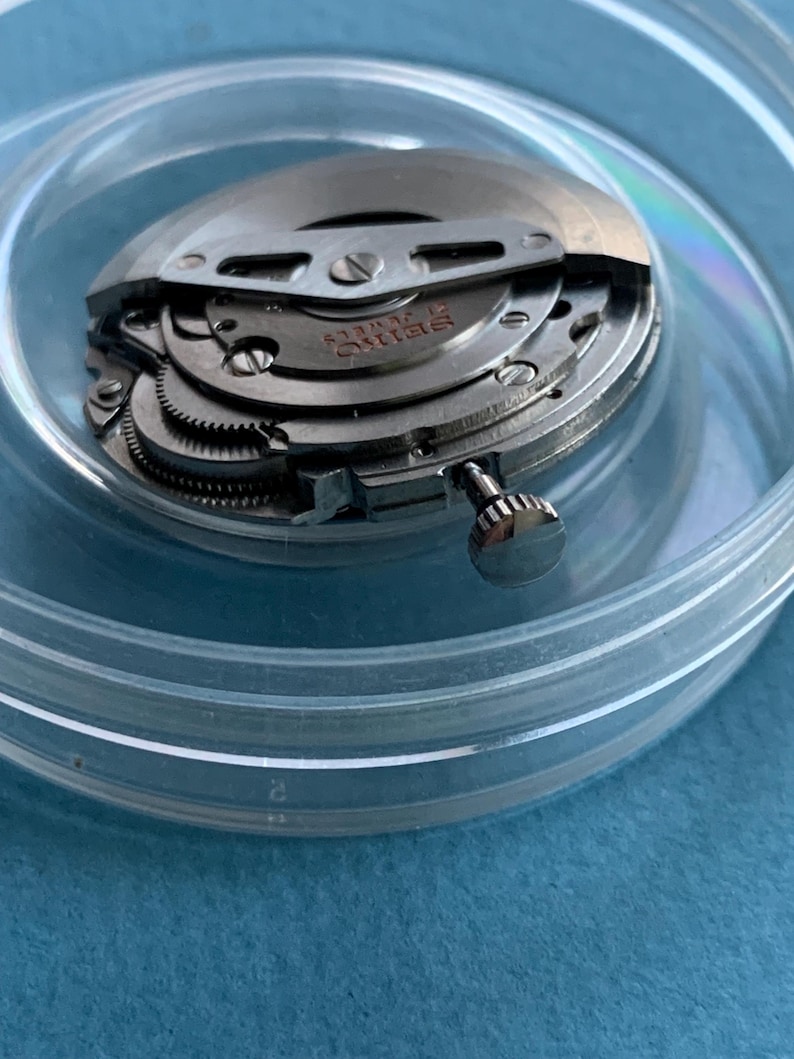 May include: A detailed view of a Seiko watch movement, revealing its complex internal mechanisms. The watch's internal parts are housed in a clear, circular container. The metal components are silver, with the word "Seiko" inscribed.