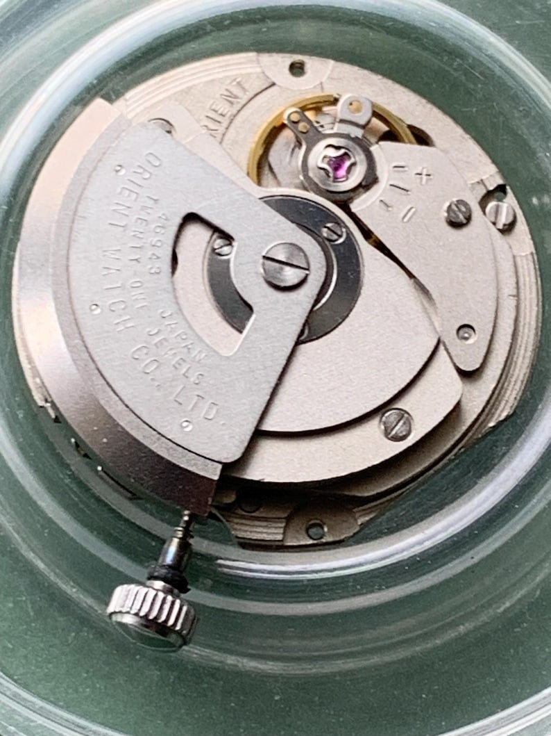 May include: Detailed view of a watch movement with silver-coloured gears, screws, and a balance wheel with a purple jewel. The text "ORIENT WATCH JAPAN JEWELS CO. LTD" is visible. The movement is housed in a clear, circular container. A small, silver crown is also visible.