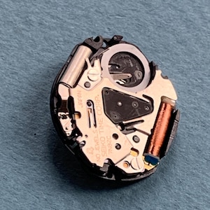 K&ouml;nnte beinhalten: Nahaufnahme eines Uhrwerks mit komplexen mechanischen Komponenten. Das Uhrwerk hat eine goldfarbene Basis mit der Aufschrift "SEIKO TIME CORP." und "JAPAN". Verschiedene Zahnr&auml;der, Spulen und andere Teile sind sichtbar und zeigen die inneren Funktionen der Uhr.