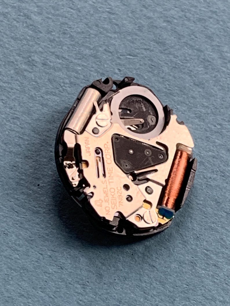 K&ouml;nnte beinhalten: Nahaufnahme eines Uhrwerks mit komplexen mechanischen Komponenten. Das Uhrwerk hat eine goldfarbene Basis mit der Aufschrift "SEIKO TIME CORP." und "JAPAN". Verschiedene Zahnr&auml;der, Spulen und andere Teile sind sichtbar und zeigen die inneren Funktionen der Uhr.