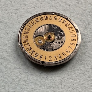 May include: Close-up of a watch movement with a gold-colored dial and silver-colored casing. The dial features numbers from 1 to 31, indicating the days of the month. The intricate gears and mechanisms are visible within the watch component.
