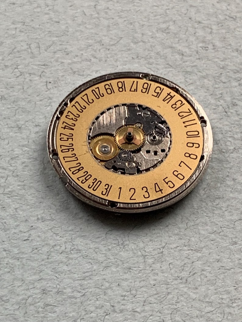 May include: Close-up of a watch movement with a gold-colored dial and silver-colored casing. The dial features numbers from 1 to 31, indicating the days of the month. The intricate gears and mechanisms are visible within the watch component.