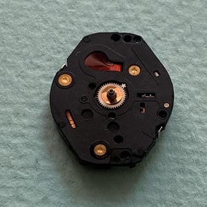 May include: A detailed view of a watch movement, showcasing a black base with gold-coloured fixings and a small, intricate gear system. A red component is visible. The watch mechanism is set against a light blue-green surface.