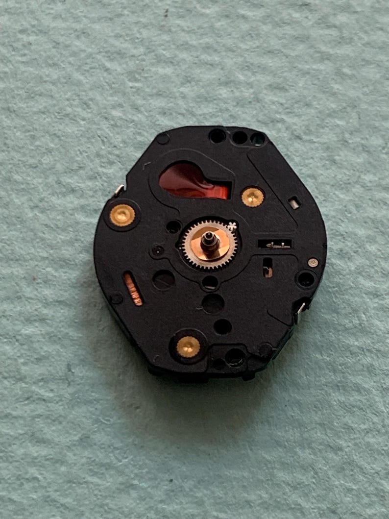 May include: A detailed view of a watch movement, showcasing a black base with gold-coloured fixings and a small, intricate gear system. A red component is visible. The watch mechanism is set against a light blue-green surface.