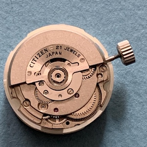 May include: A Citizen watch movement with the text "Citizen Japan 21 Jewels". The watch mechanism is silver and beige, with intricate gears and a winding stem. The watch movement is circular.
