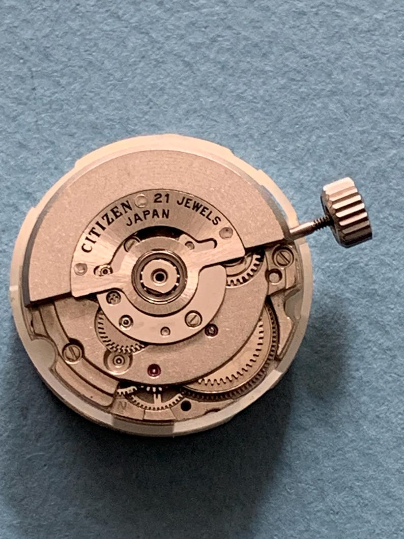 May include: A Citizen watch movement with the text "Citizen Japan 21 Jewels". The watch mechanism is silver and beige, with intricate gears and a winding stem. The watch movement is circular.