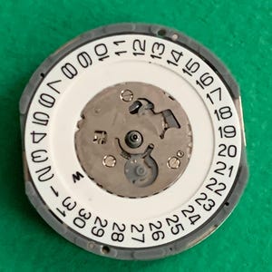May include: A watch movement component with a white date dial featuring black numbers from 1 to 31. The central mechanism is silver-colored, surrounded by a gray outer ring. The component is set against a green background.