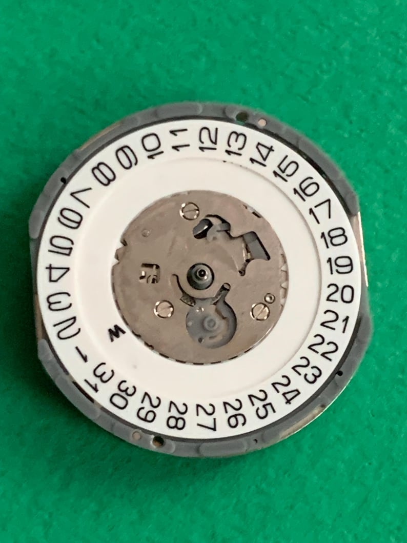 May include: A watch movement component with a white date dial featuring black numbers from 1 to 31. The central mechanism is silver-colored, surrounded by a gray outer ring. The component is set against a green background.