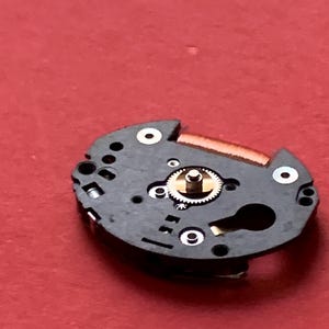 May include: Close-up of a watch movement component. The black, circular piece has several holes and a small, gold-colored gear in the center. A thin, copper-colored piece is attached to the top. The background is a solid red.