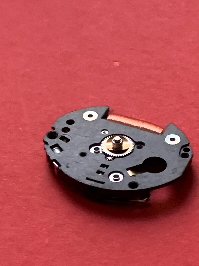 May include: Close-up of a watch movement component. The black, circular piece has several holes and a small, gold-colored gear in the center. A thin, copper-colored piece is attached to the top. The background is a solid red.