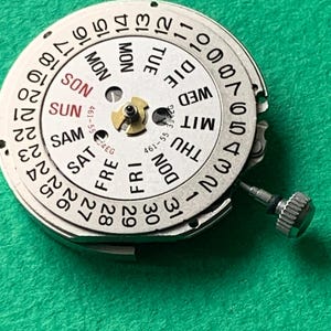 May include: A close-up of a watch movement with a white dial and black numerals. The dial displays the days of the week in English and the numbers 1-31. The text "461-55" is visible. A small silver crown is attached.
