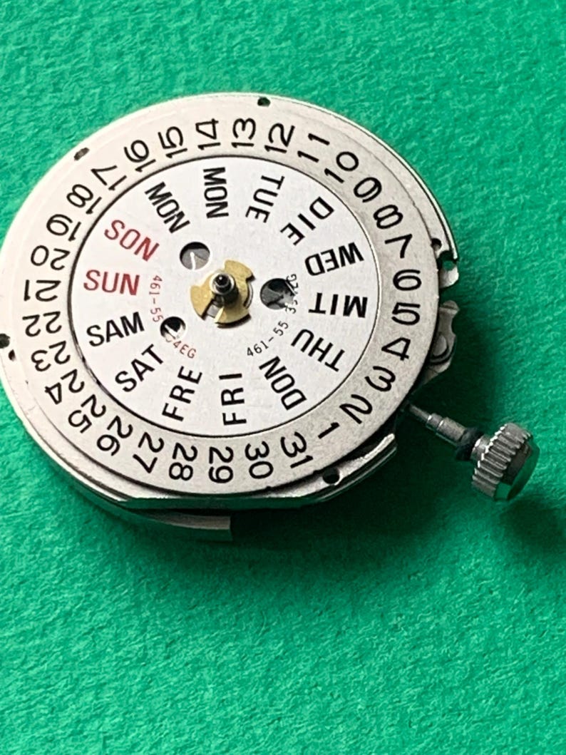 May include: A close-up of a watch movement with a white dial and black numerals. The dial displays the days of the week in English and the numbers 1-31. The text "461-55" is visible. A small silver crown is attached.