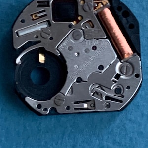 May include: Close-up of a watch movement with silver and gold components. The movement has a black, octagonal frame and a copper coil. The text "ORIENT WATCH CO LTD" and "JAPAN" are visible.