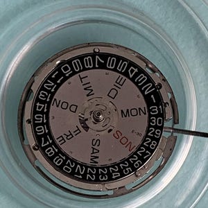 May include: Close-up of a watch movement with a silver dial and black numerals. The dial displays the days of the week and the date, with the numbers 1-31. The words "SAM", "DON", "FRE", "MON", and "DIE" are visible.