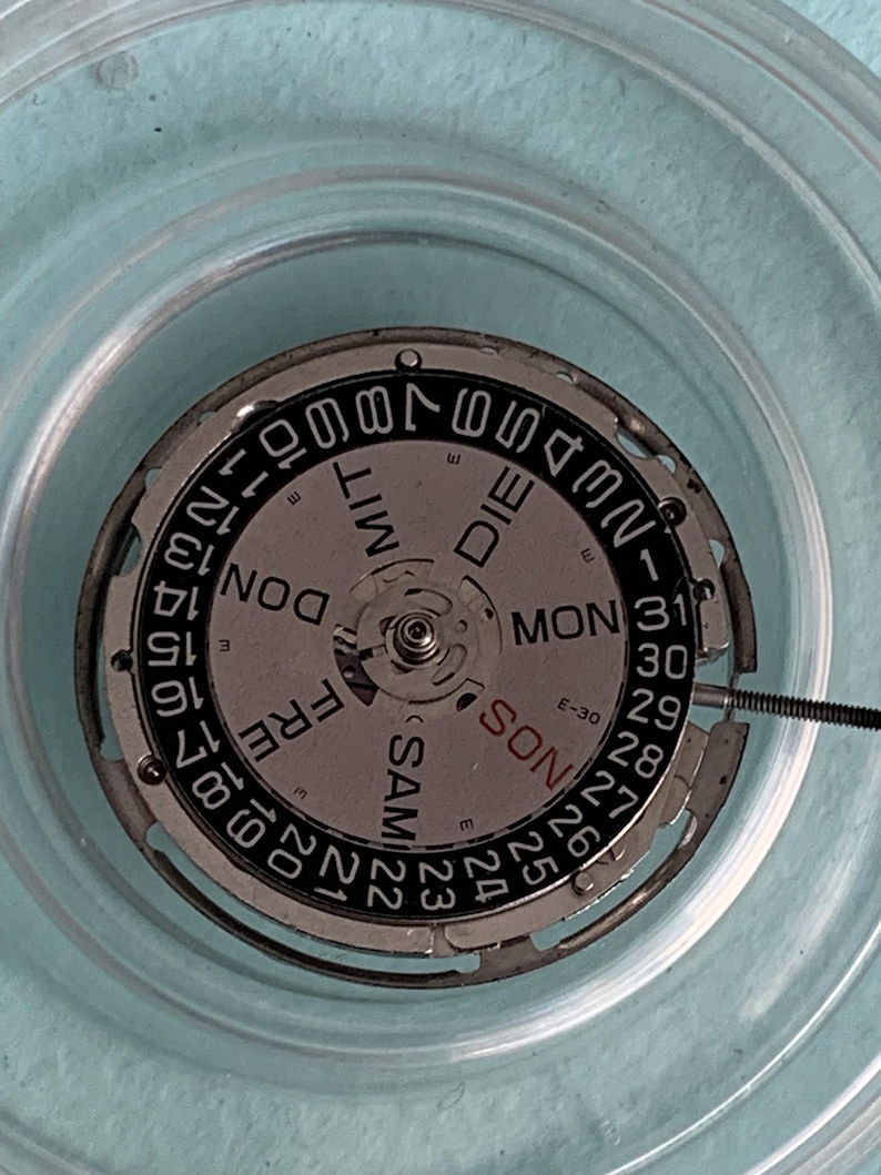 May include: Close-up of a watch movement with a silver dial and black numerals. The dial displays the days of the week and the date, with the numbers 1-31. The words "SAM", "DON", "FRE", "MON", and "DIE" are visible.
