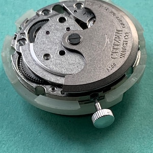 May include: A close-up of a Citizen watch movement, featuring intricate gears and components. The watch mechanism is silver-toned with the text "CITIZEN WATCH CO." and "21 JEWELS" visible. The watch movement is encased in a white plastic frame.