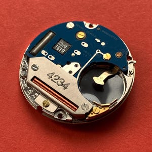 May include: A close-up of a watch movement with a blue circuit board and silver metal components. The text "SEVEN 7 JEWELS" and "UNADJUSTED" are visible. The number "4234" is printed on the metal.