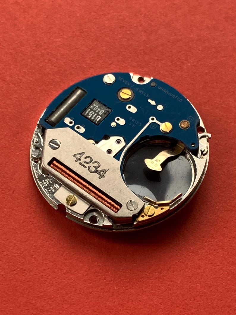 May include: A close-up of a watch movement with a blue circuit board and silver metal components. The text "SEVEN 7 JEWELS" and "UNADJUSTED" are visible. The number "4234" is printed on the metal.