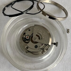 May include: A disassembled watch movement with various components. The main mechanism is silver-toned, with intricate gears and a red jewel. Black rubber rings and a silver metal ring are also present, along with small silver-colored ball bearings.