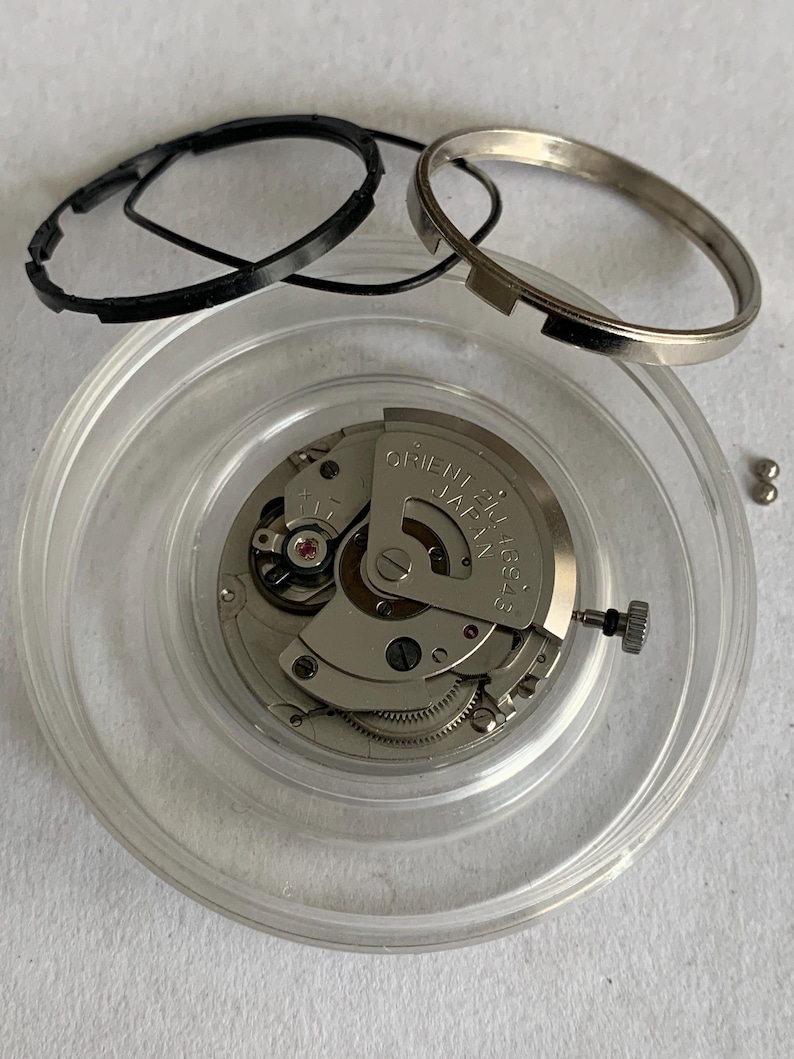May include: A disassembled watch movement with various components. The main mechanism is silver-toned, with intricate gears and a red jewel. Black rubber rings and a silver metal ring are also present, along with small silver-colored ball bearings.