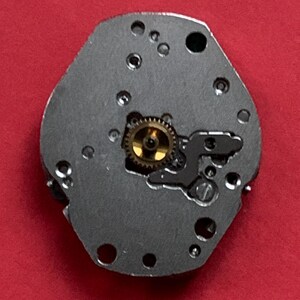 May include: Close-up of a watch movement component. The component is a silver-colored, irregularly shaped plate with several small holes and a central gold-colored gear. The background is a solid red color.