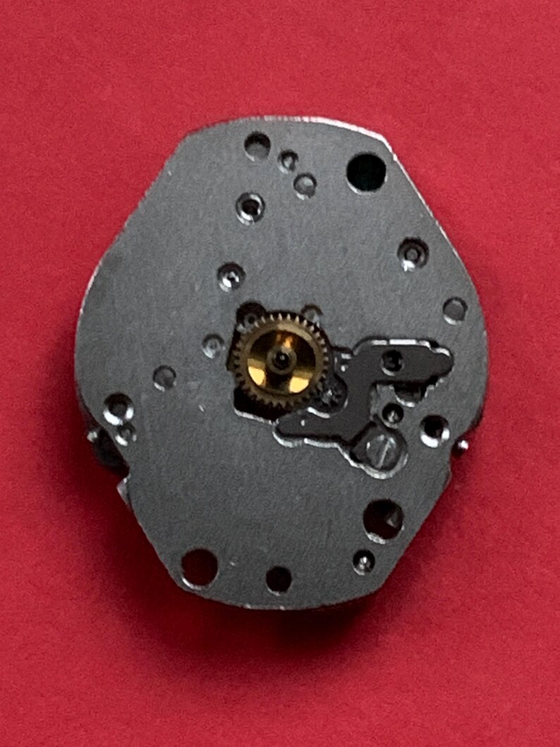 May include: Close-up of a watch movement component. The component is a silver-colored, irregularly shaped plate with several small holes and a central gold-colored gear. The background is a solid red color.