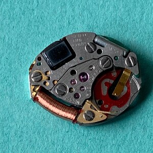 May include: Close-up of a watch movement with various gears, screws, and a red rotor. The movement is silver with gold accents and a copper coil. The text "SEIKO TIME CORP" and "JAPAN" are visible.