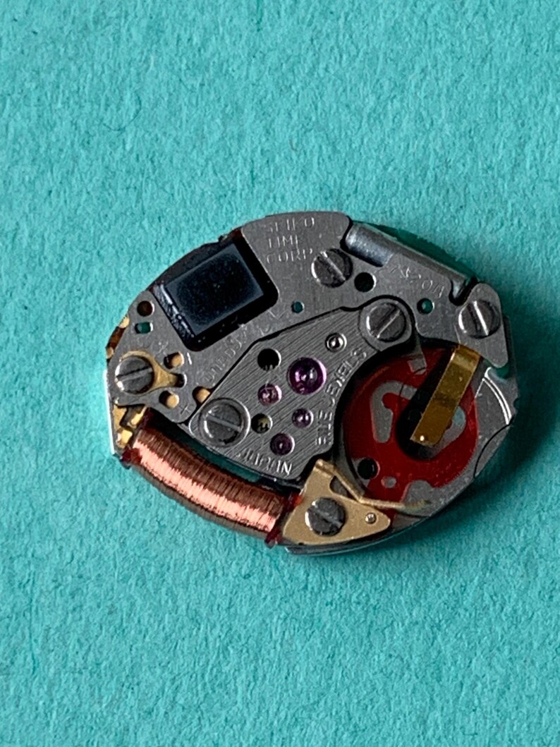 May include: Close-up of a watch movement with various gears, screws, and a red rotor. The movement is silver with gold accents and a copper coil. The text "SEIKO TIME CORP" and "JAPAN" are visible.
