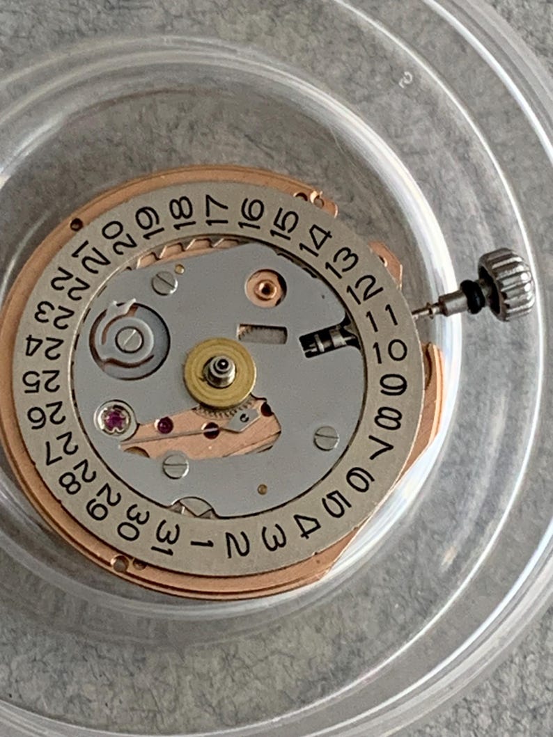 Puede incluir: Primer plano de un movimiento de reloj con una esfera plateada y dorada. La esfera presenta n&uacute;meros negros para la fecha, del 1 al 31. Se ve una peque&ntilde;a corona plateada, junto con varios engranajes y joyas. El movimiento est&aacute; dentro de un recipiente de pl&aacute;stico transparente.